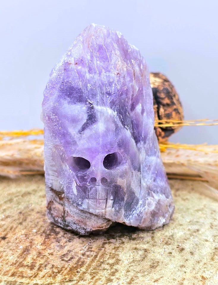 A raw purple amethyst crystal skull with natural striations and rough edges, displayed on a textured beige stone surface with dried grass and a blurred background.