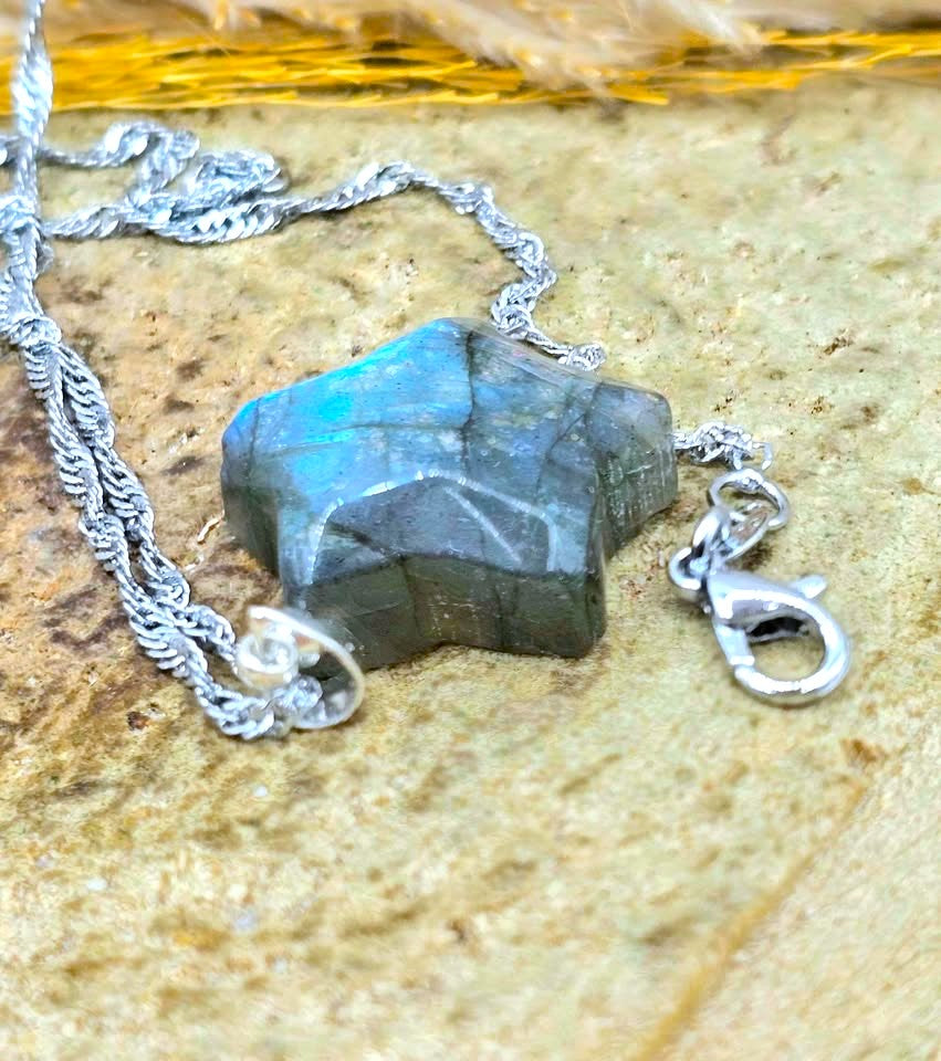 A silver chain necklace featuring a faceted dark gray labradorite gemstone pendant with a blue-green flash, resting on a textured beige stone surface.