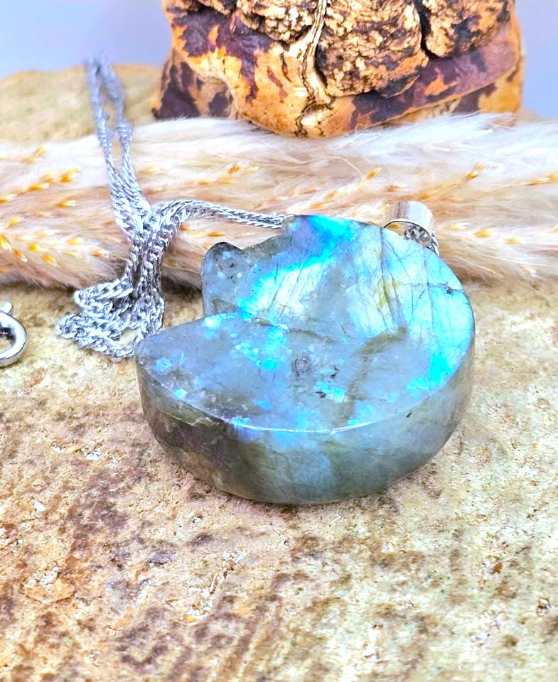 A cat-shaped labradorite pendant necklace with a silver chain, resting on a textured stone surface with dried grass and a bark piece in the background.