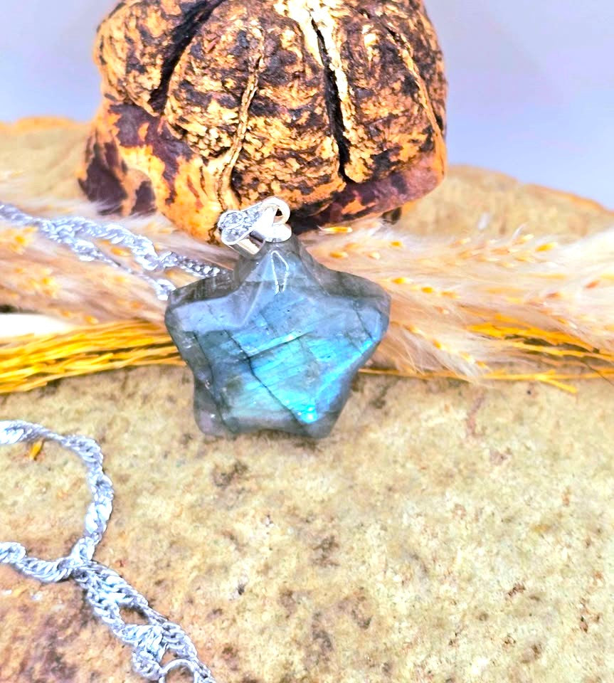 A star-shaped labradorite gemstone pendant necklace with a silver bail and chain, displayed on a textured stone surface with dried wheat and a decorative object in the background.