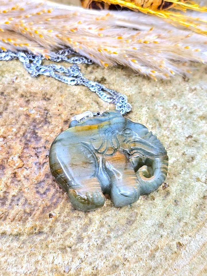 A carved elephant pendant necklace made of polished gray stone with natural brown and greenish veining, hanging from a silver-toned chain on a textured stone surface with dried grass in the background.