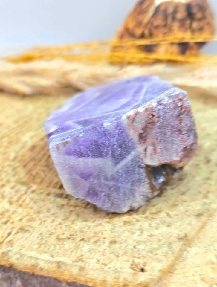 A raw purple amethyst crystal with rough, natural facets resting on a textured beige stone slab.