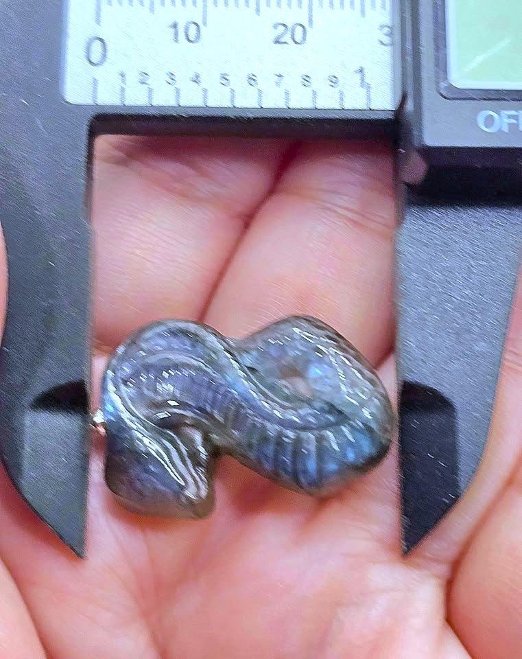 A small polished gray stone or bead held in a person's hand, with a digital caliper above it for size reference.