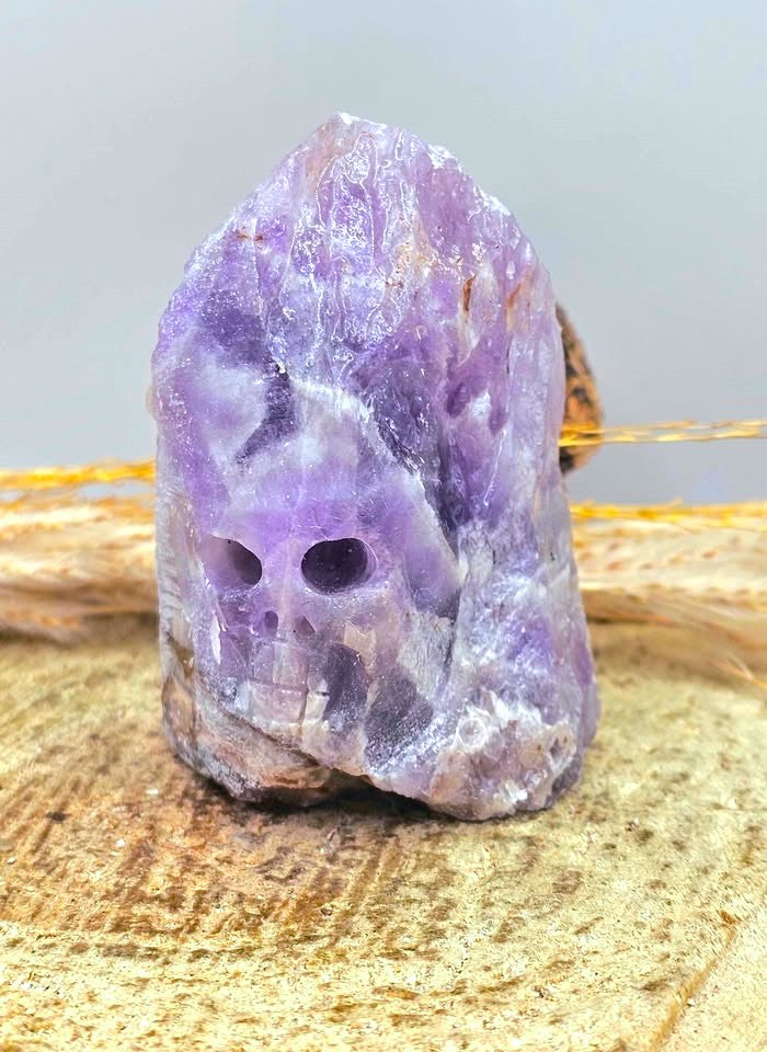 A raw purple amethyst crystal skull with natural rough edges, resting on a textured wooden surface with dried grass in the background.