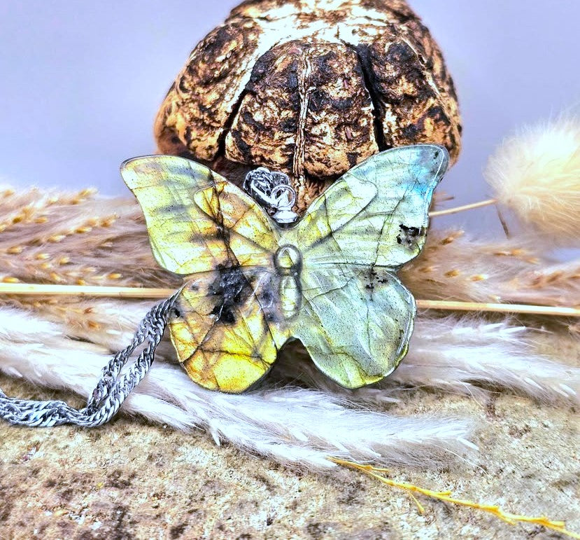 A butterfly-shaped pendant with a silver-toned chain, featuring yellow, green, and black marbled wings, displayed on a textured surface with dried grass and a brown decorative object in the background.