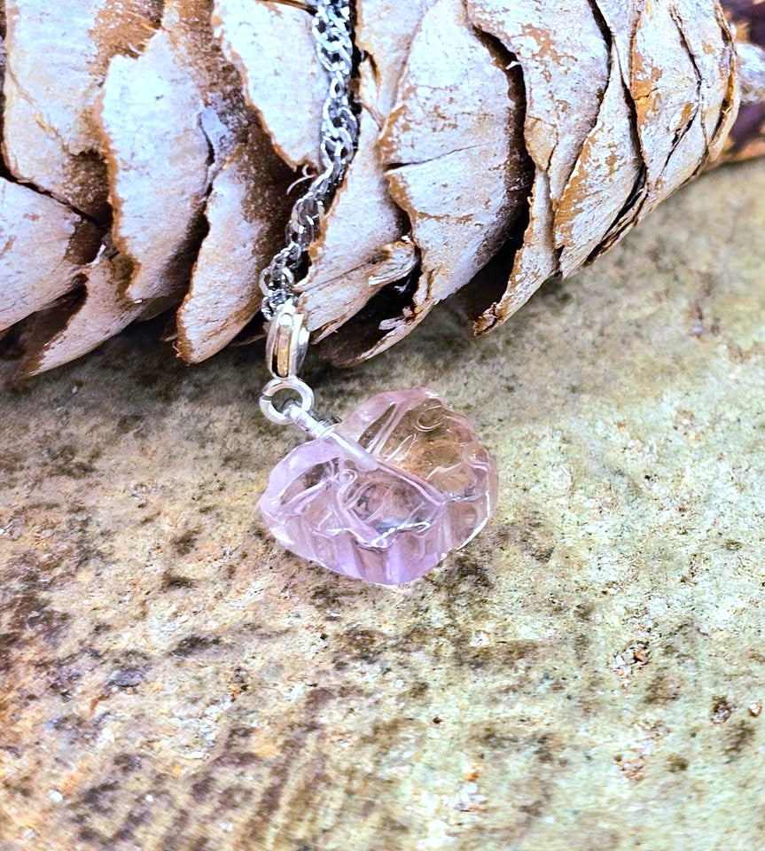 A close-up of a silver chain necklace with a faceted, organic-shaped light pink crystal pendant resting on a textured stone surface next to a pine cone.
