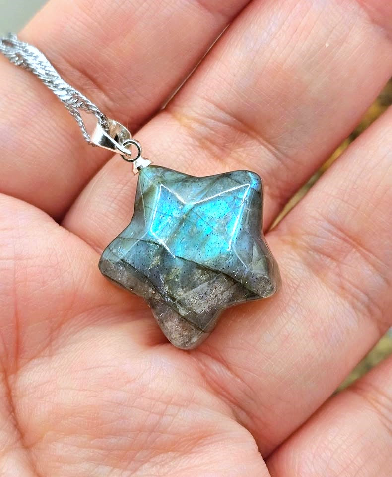 A faceted star-shaped labradorite gemstone pendant on a silver chain, held in a person's hand to show scale.