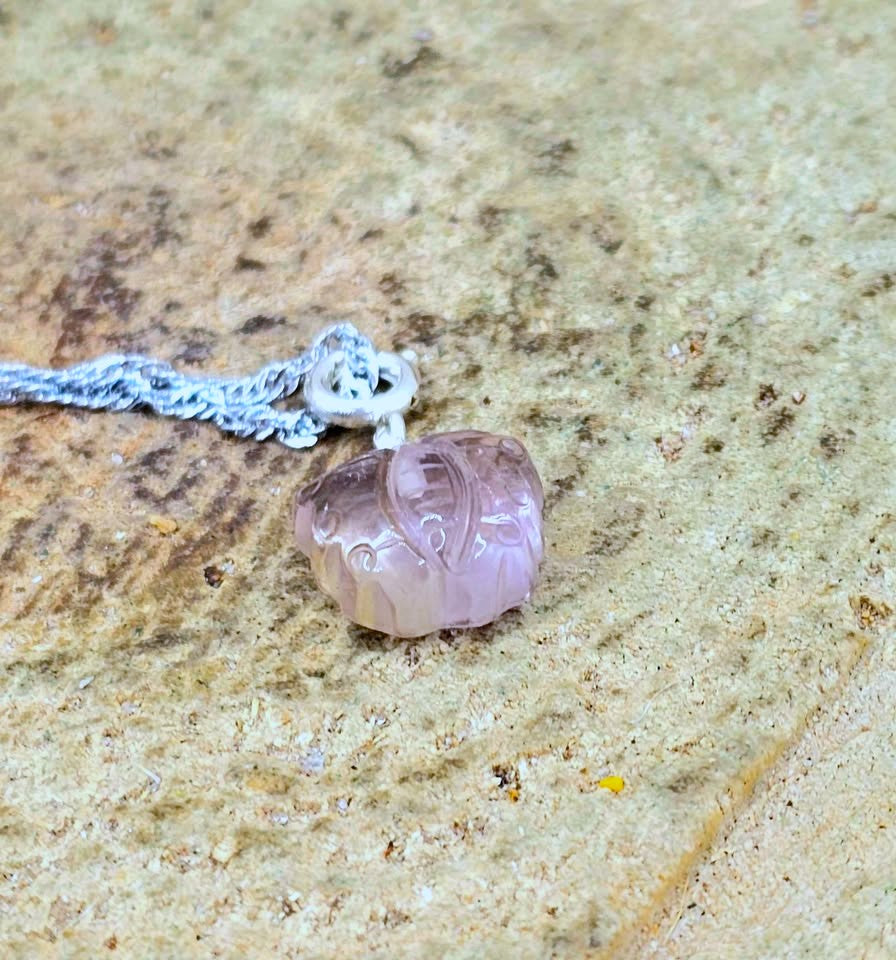 A carved light purple lotus pendant necklace with a silver bail and silver chain, resting on a textured beige stone surface.