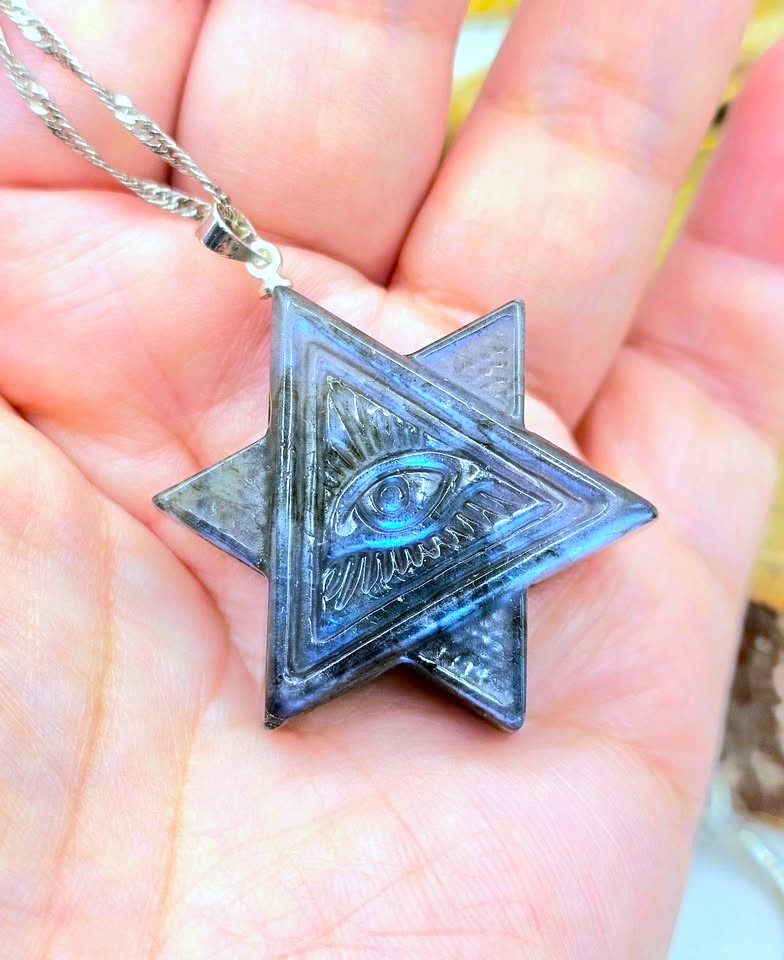 A close-up of a silver-toned Star of David pendant with an eye of providence design, held in a person's palm. The pendant has a textured, metallic finish with a silver chain visible at the top left.