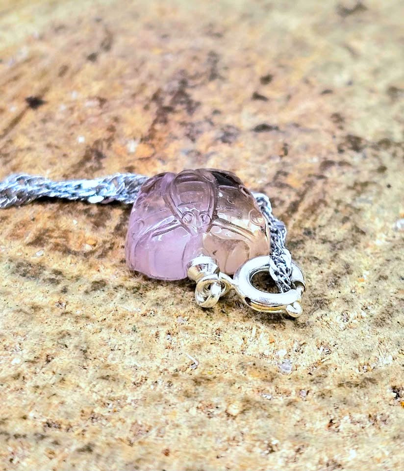 A close-up of a silver chain necklace with a carved light purple amethyst beetle pendant resting on a textured brown stone surface.