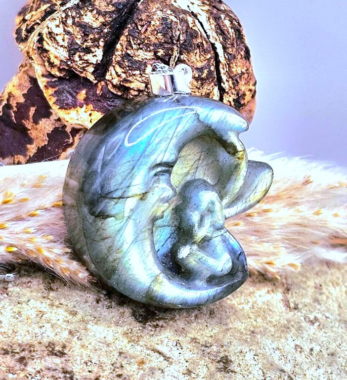 A polished labradorite crescent moon pendant with a carved sleeping figure, displayed on a textured surface with a fur pelt and a brown shell in the background.