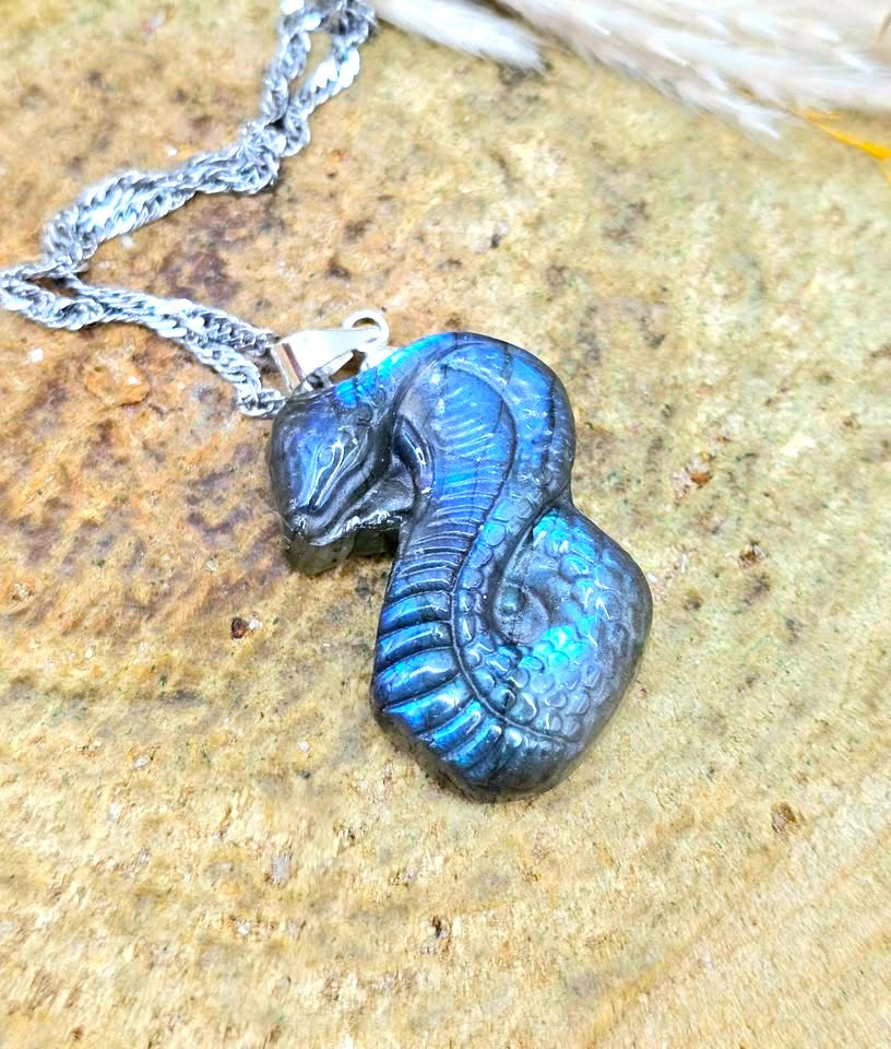 A carved snake pendant necklace with a textured blue and black finish, resting on a natural stone surface with a silver chain.