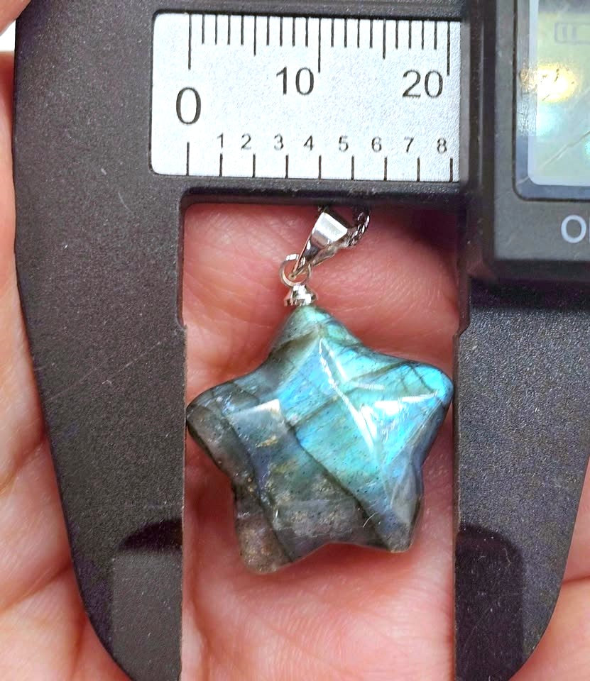 A star-shaped labradorite gemstone pendant held in a hand under a metal caliper, showing its blue-green iridescent color shift and silver bail.