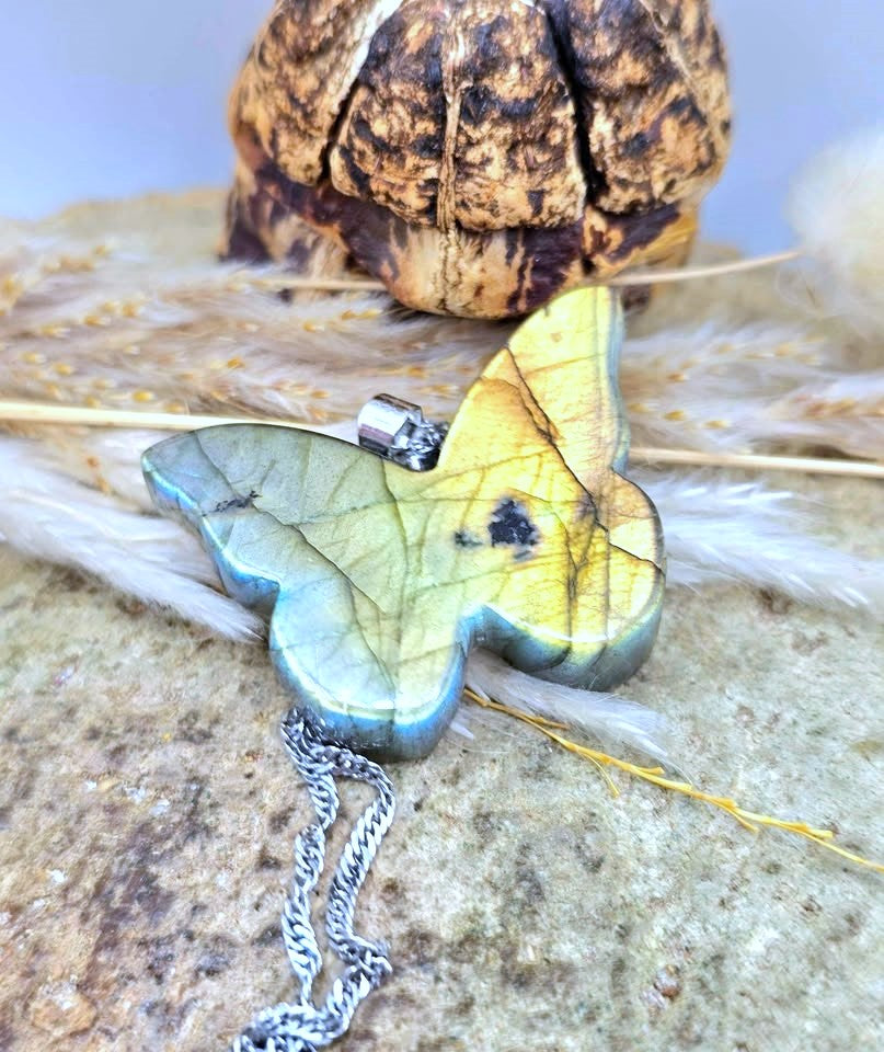 A butterfly-shaped pendant necklace made from iridescent labradorite stone, resting on a textured stone surface with dried grass and a decorative object in the background. The pendant shows a yellow, green, and blue color shift with a silver-toned chain.