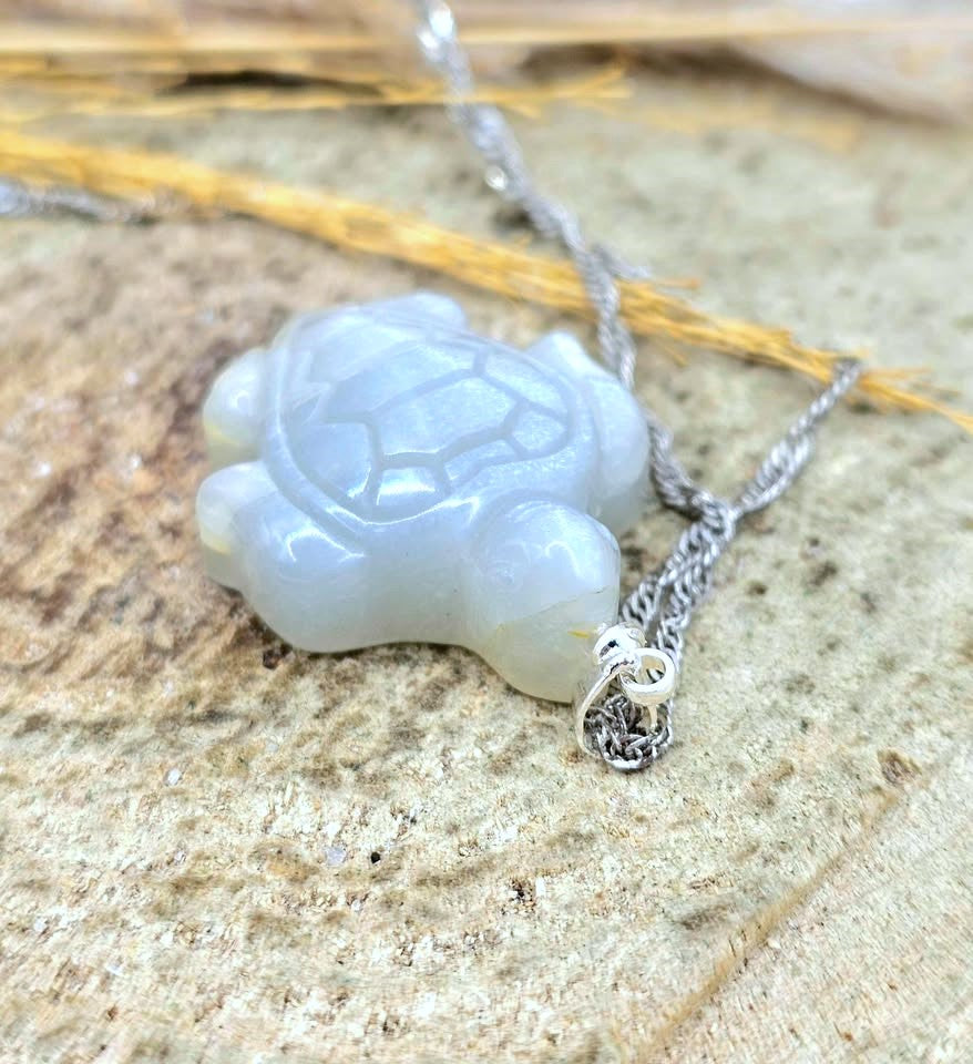 A carved turtle pendant necklace in a pale milky white stone, attached to a silver chain, resting on a textured stone surface with dried grass.