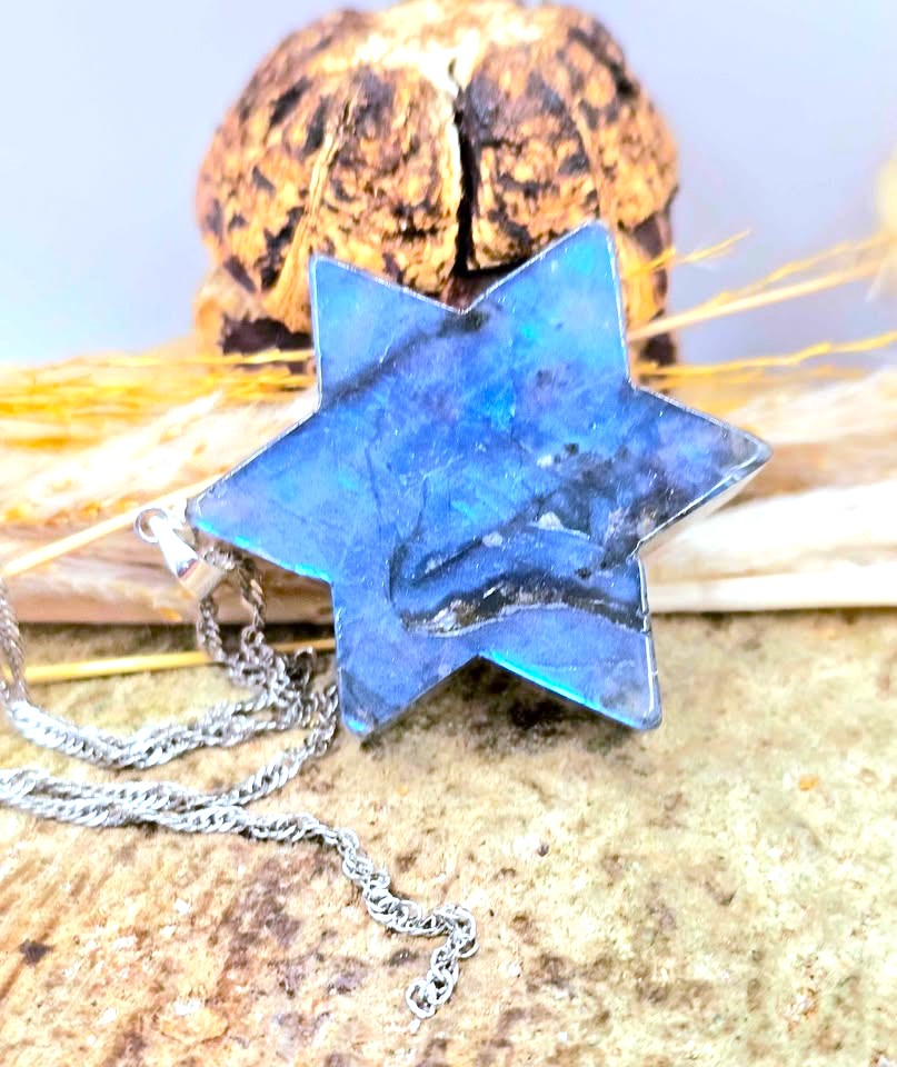 A six-pointed star pendant made of polished blue labradorite with a silver-toned chain, displayed on a textured stone surface with dried grass and a decorative object in the background.