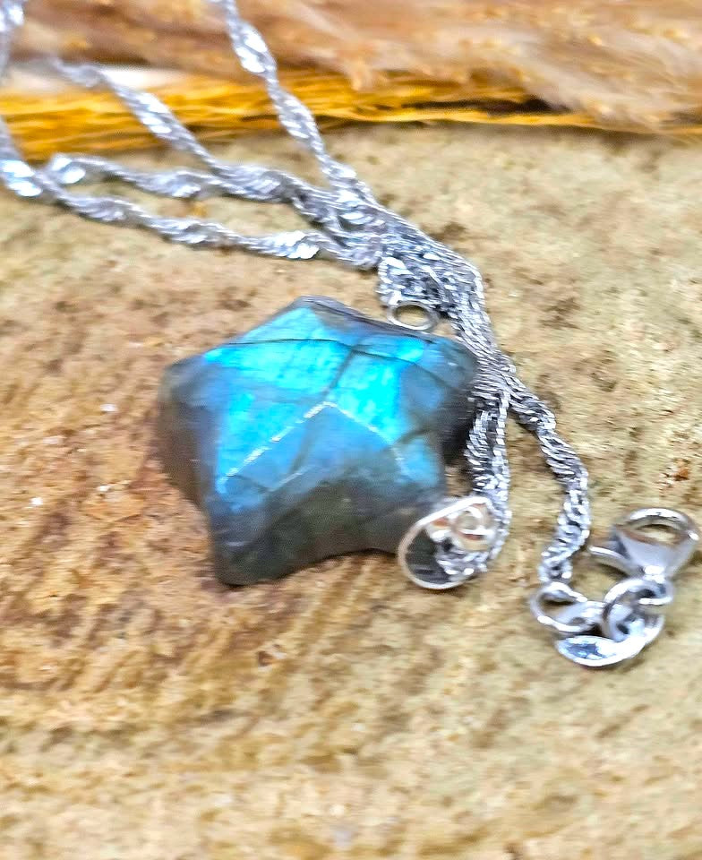 A silver chain necklace with a faceted, irregular dark gray-green stone pendant showing vibrant blue iridescent flashes, resting on a textured tan surface.