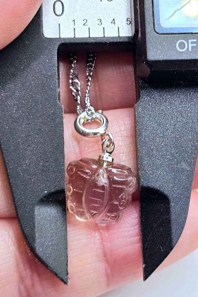 A close-up of a small pendant necklace held in a digital caliper. The pendant appears to be a clear faceted bead or charm with a silver-toned chain and clasp.