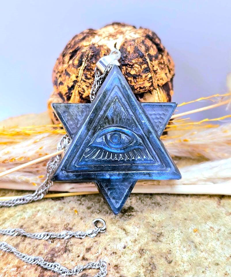 A dark blue labradorite pyramids pendant with an all-seeing eye design, hanging from a silver chain, styled on a textured stone surface with dried grass and a decorative object in the background.