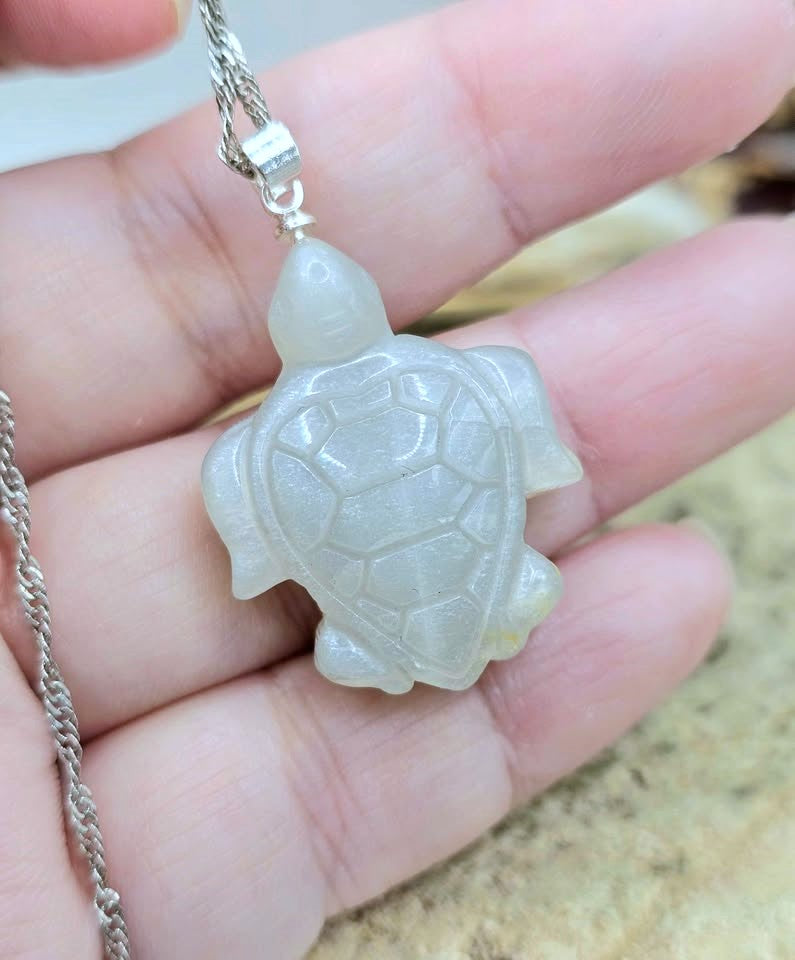 A hand holding a carved turtle pendant necklace made of milky translucent stone, attached to a silver-tone chain.