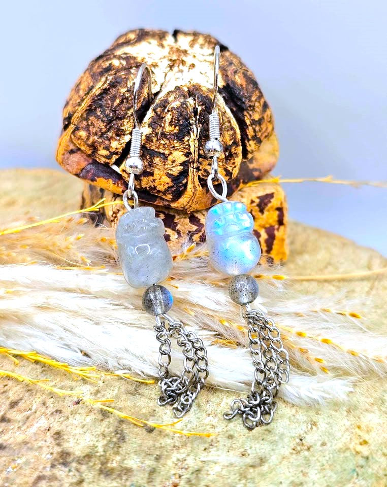 A pair of bohemian-style dangle earrings displayed on a textured rock with dried grass. The earrings feature silver-toned chains, small gray beads, and light blue iridescent stone beads (labradorite) with carved silver Buddha charms.