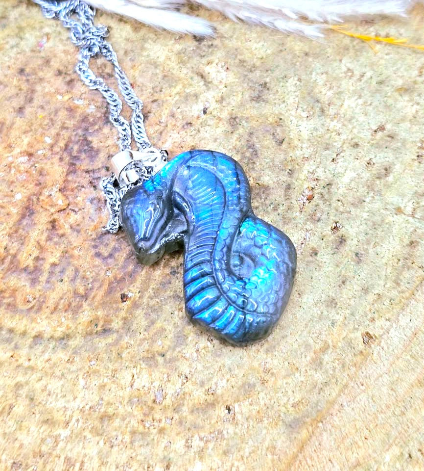 A silver chain necklace with a sculpted snake pendant featuring a blue and green iridescent finish, resting on a textured stone surface.