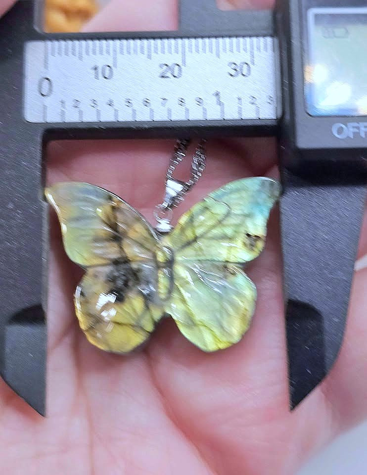 A butterfly-shaped pendant with iridescent green and yellow wings held in a hand under a caliper to show its size. The pendant is attached to a silver chain.