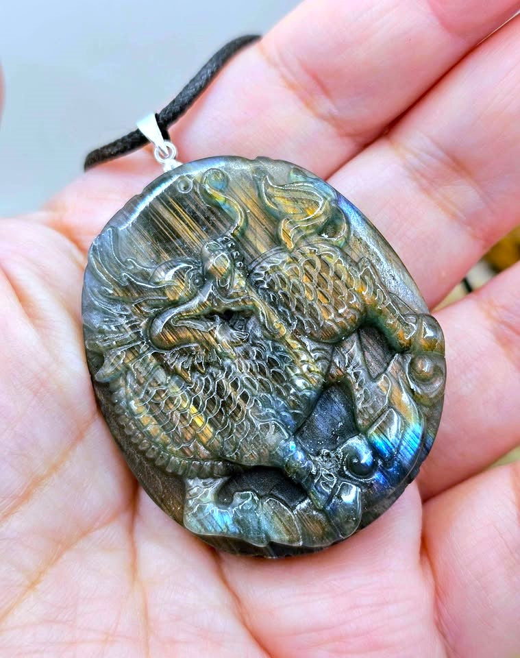 A hand holding an oval carved pendant made of iridescent labradorite stone with a raised dragon design, attached to a black cord.