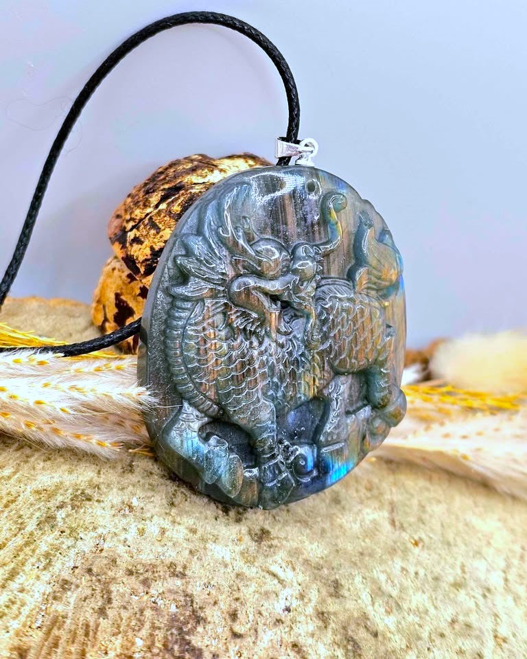 A carved oval pendant necklace featuring a detailed dragon design, made from a dark gray labradorite stone with blue-green iridescent flashes. The pendant is suspended from a black cord and is displayed on a textured beige surface with a decorative shell in the background.