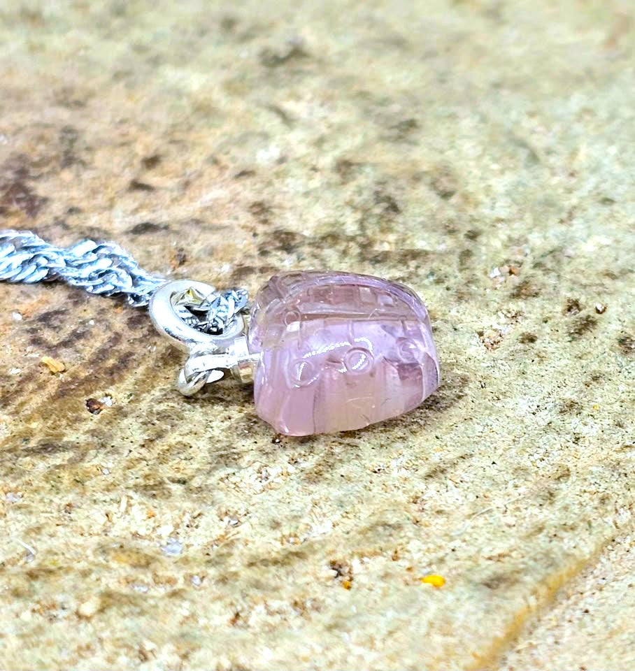 A close-up of a carved light purple amethyst pendant on a silver chain, resting on a textured beige stone surface.