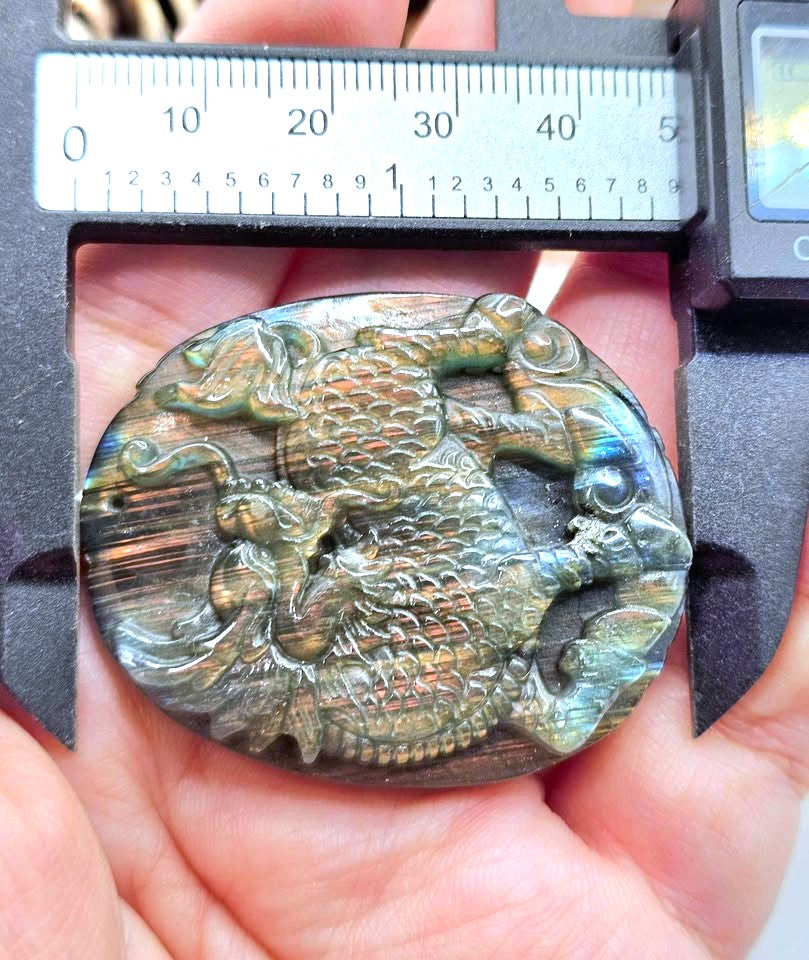 A close-up of a hand holding an oval iridescent stone carving with a raised dragon motif, with a digital caliper measuring the top and bottom edges for scale.