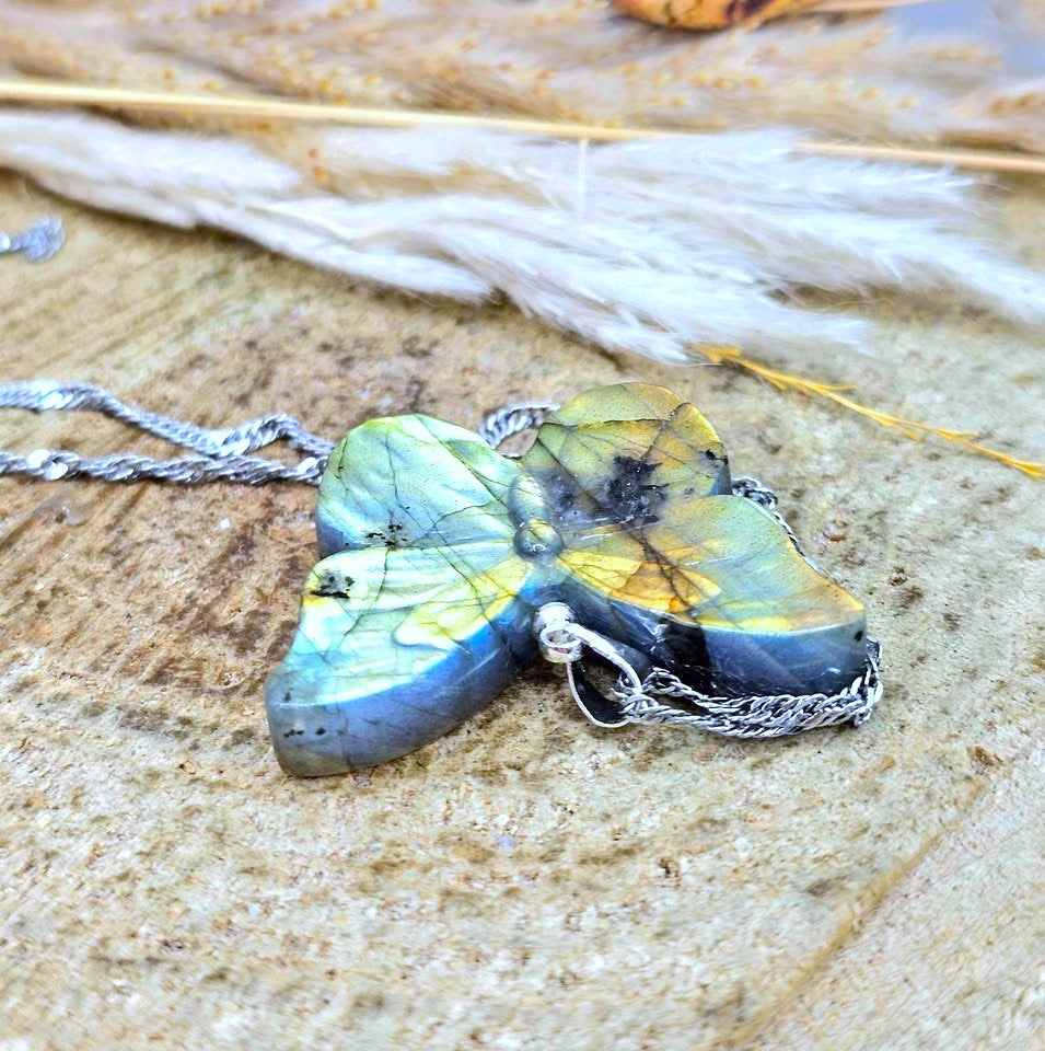 A butterfly-shaped pendant necklace made from iridescent labradorite stone with silver chain, resting on a textured stone surface with dried grass in the background.