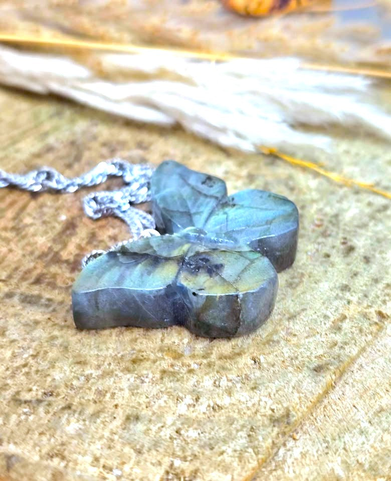 A close-up of a raw labradorite stone pendant on a silver chain, resting on a textured stone surface with dried grass in the background.