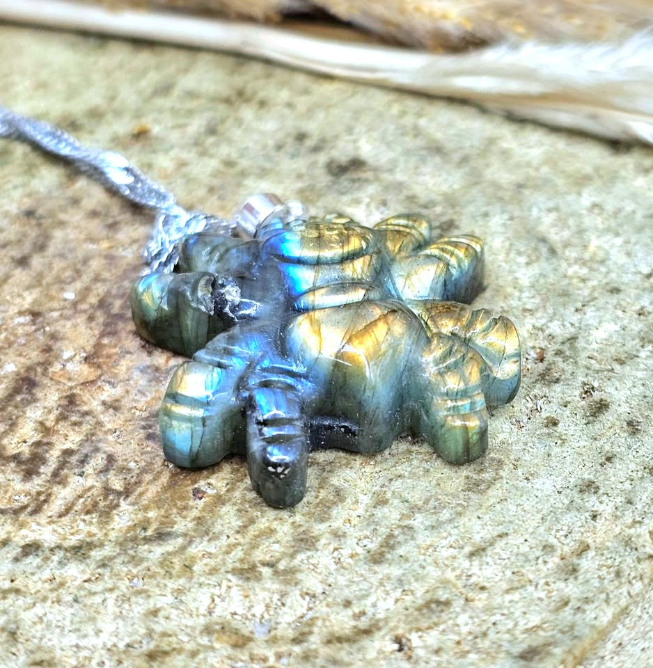 A small carved pendant or charm with an iridescent blue-green-gold labradorite-like finish, attached to a silver cord, resting on a textured stone surface.