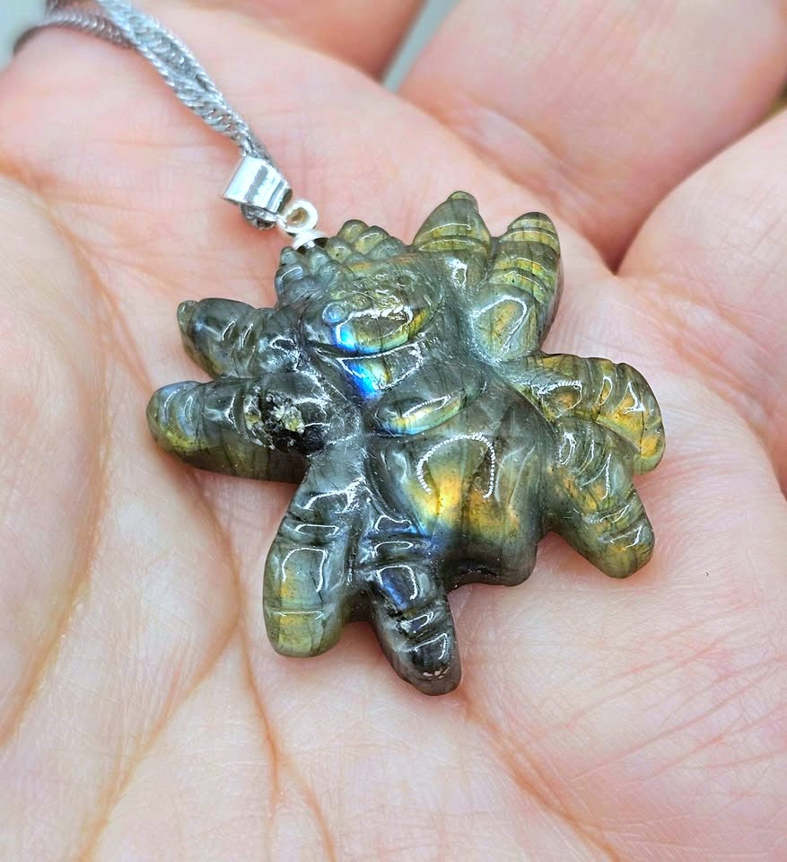 A carved labradorite pendant shaped like a five-pointed star or sunburst, resting on a person's palm. The pendant shows natural green and gold iridescent labradorite flashes and is suspended from a silver chain.