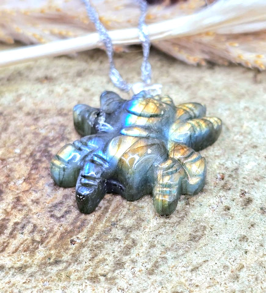 A carved labradorite gemstone pendant shaped like a flower or starburst, hanging from a silver-toned chain against a textured stone surface with dried leaves in the background.