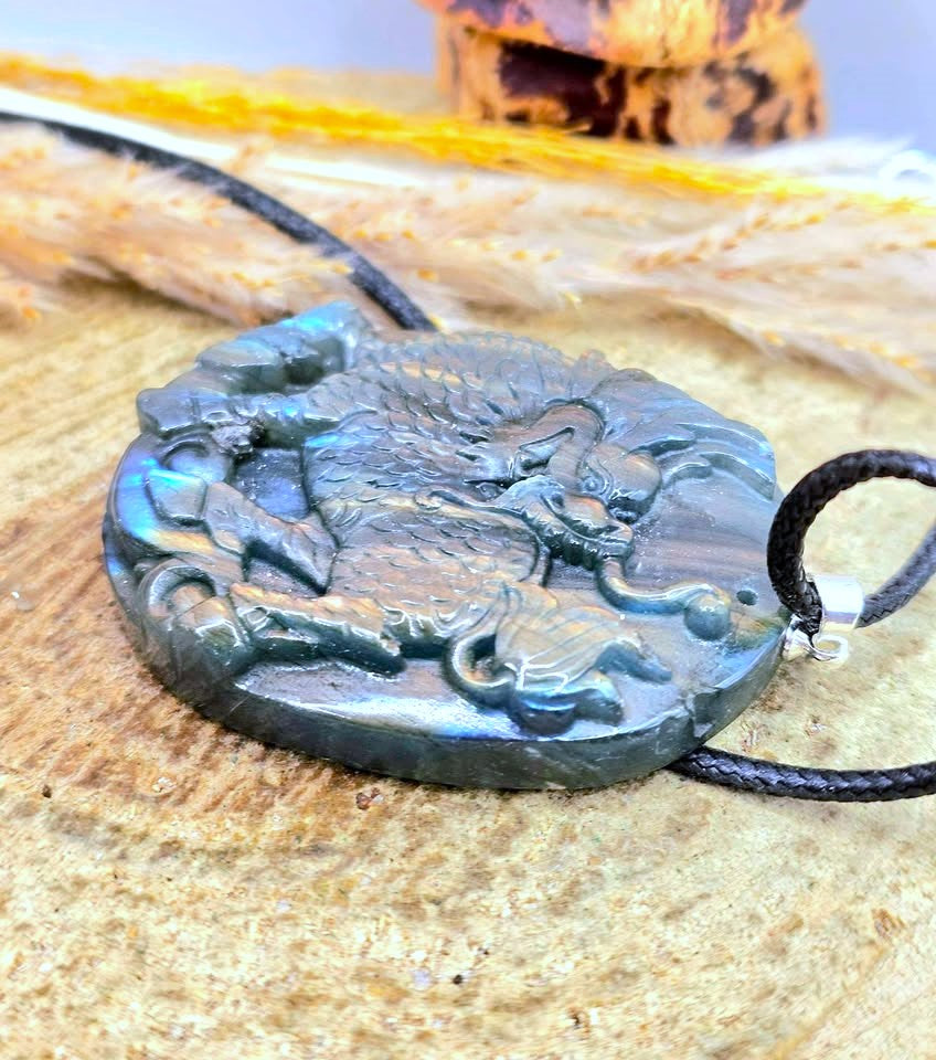 A carved oval pendant necklace featuring a dark dragon design with iridescent blue-green highlights, suspended on a black cord over a textured wood surface.