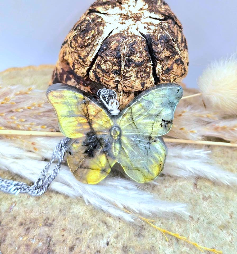A butterfly-shaped pendant necklace with yellow, green, and black marbled wings, resting on dried grass in front of a textured brown decorative object.