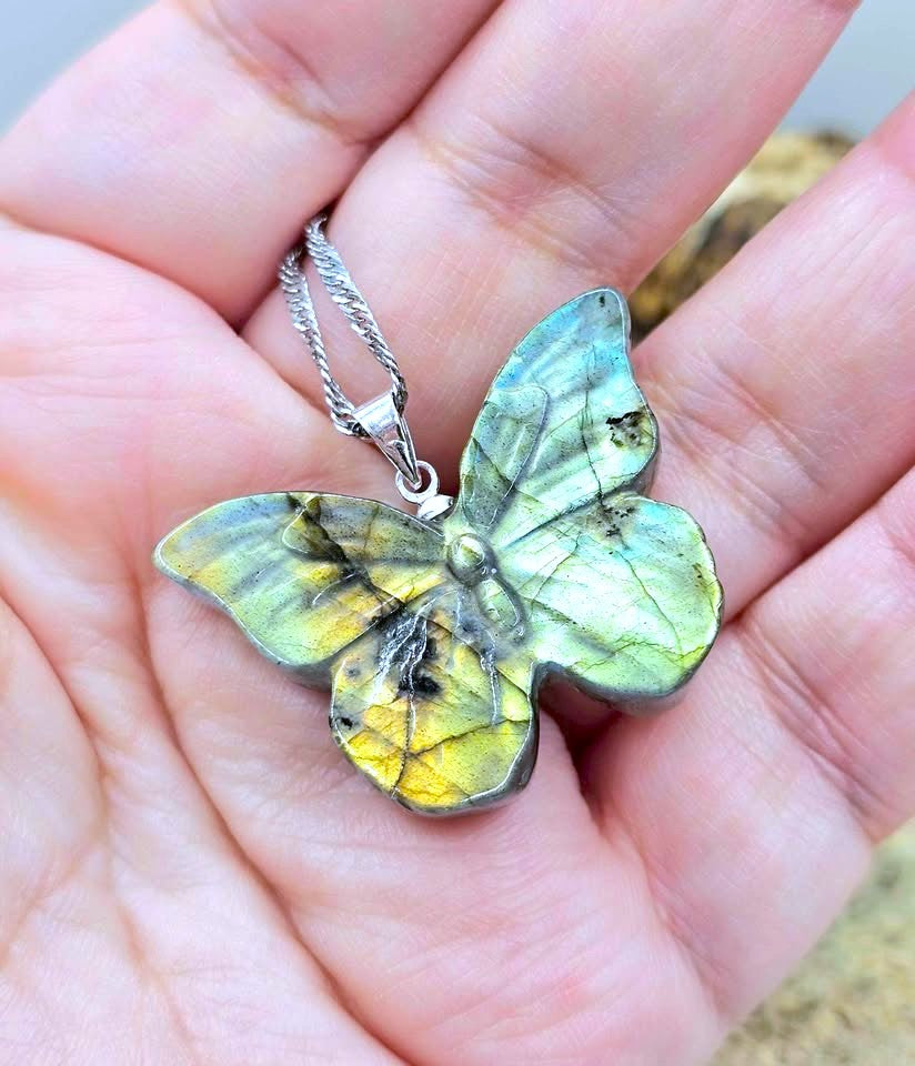 A butterfly-shaped pendant made from polished labradorite stone with a silver chain, held in a person's hand. The labradorite displays a shimmering color shift between yellow, green, and blue-gray.