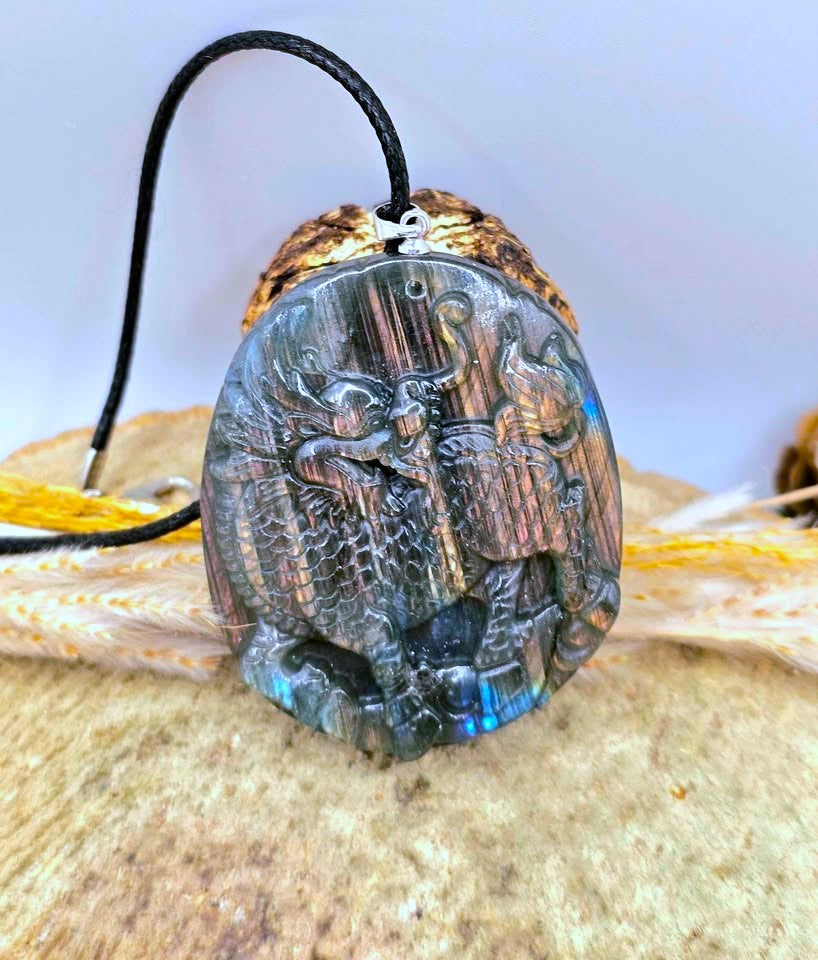 A carved oval pendant featuring a detailed relief scene of figures, made from a dark iridescent stone with blue and gold flashes, hanging from a black cord on a textured beige surface.