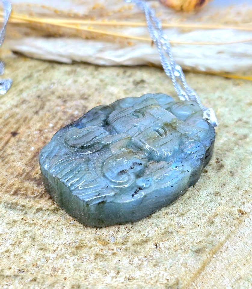 A carved stone pendant necklace with a silver-toned chain, resting on a textured sandy surface. The pendant is made of a polished gray stone with natural inclusions and a smooth, polished finish.