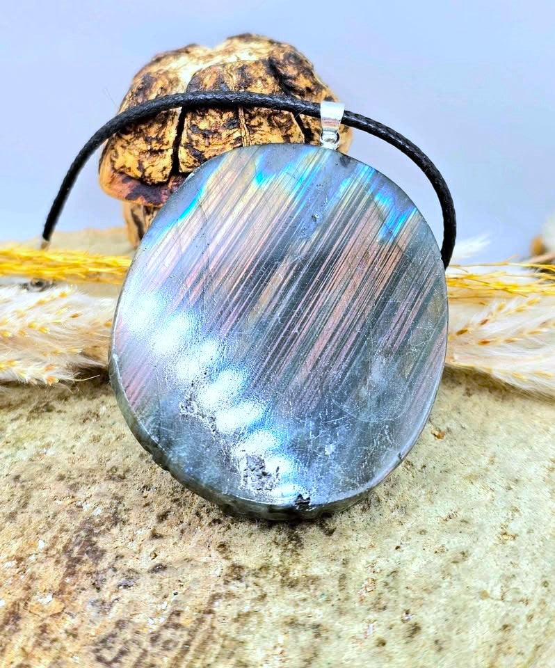 A round labradorite pendant necklace with a black cord and silver bail, resting on a textured stone surface with dried grass and a small rock in the background.