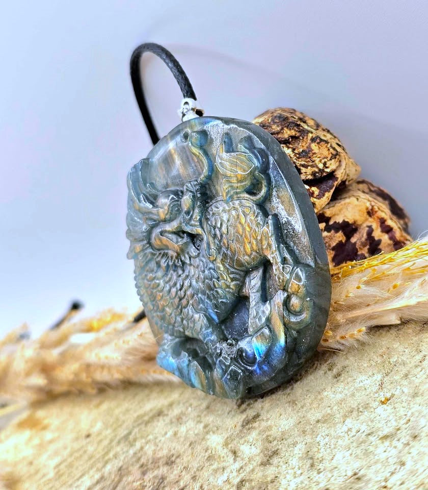 A carved oval pendant featuring a detailed dragon design in gray labradorite with iridescent blue and gold flashes, hanging from a black cord, displayed on a sandy surface with dried grass and a shell.