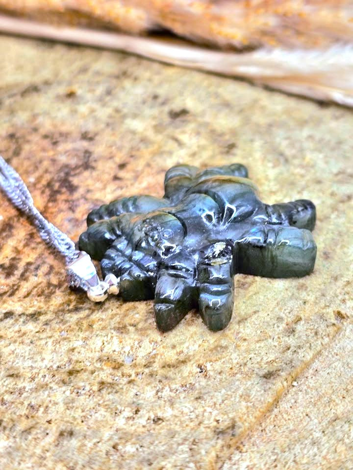 A carved black stone pendant shaped like a five-pointed star or hand, resting on a textured sandy surface with a silver-colored cord attached.
