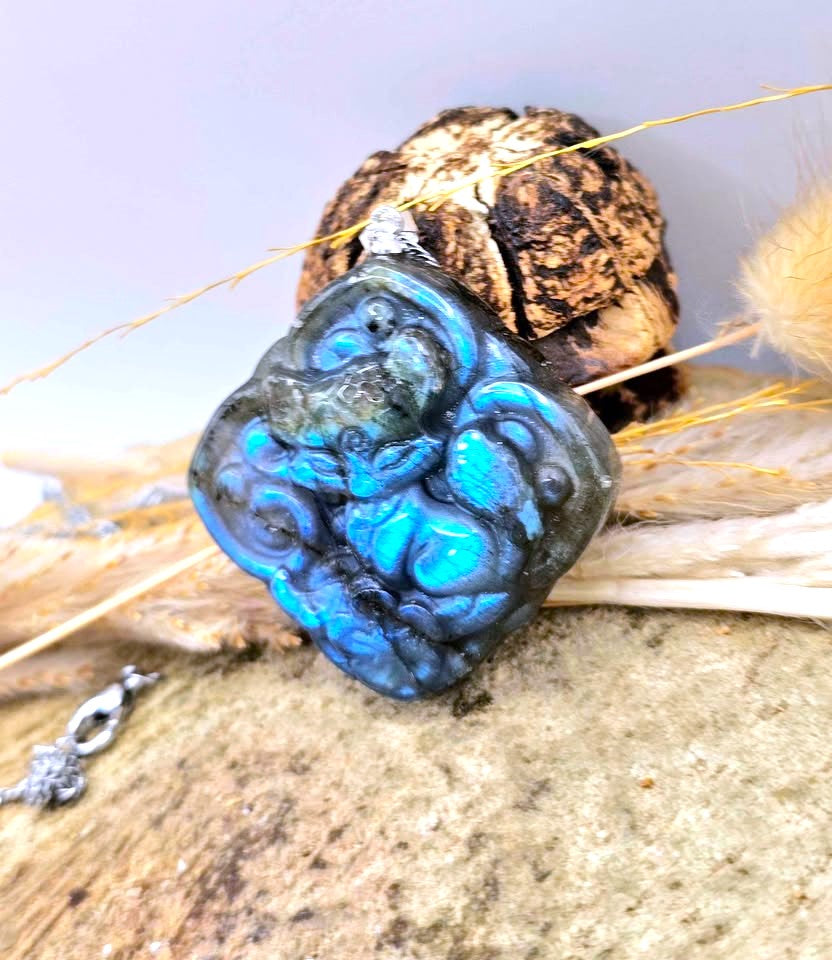 A carved blue agate pendant featuring a detailed Buddha design, displayed on a textured stone surface with dried grass and a decorative shell in the background.