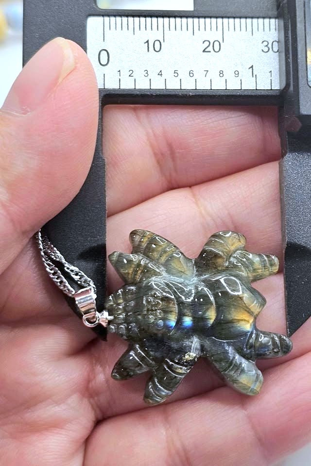 A close-up of a hand holding a small shell-shaped pendant or charm with a silver chain, positioned under a caliper for size reference.