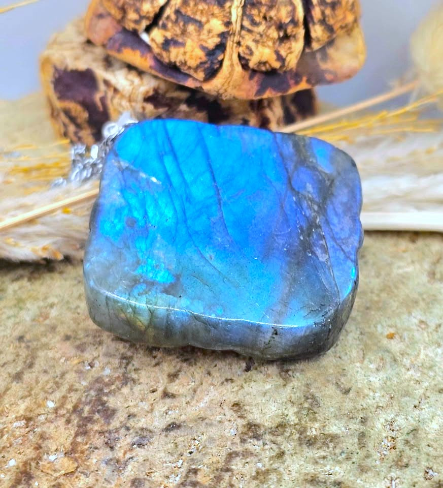 A raw labradorite gemstone pendant with vibrant blue and green iridescent flashes, resting on a textured stone surface with dried grass and a decorative object in the background.