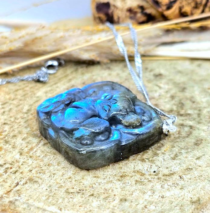 A carved Labradorite pendant necklace featuring a sleeping figure, displayed on a textured stone surface with dried grass in the background.