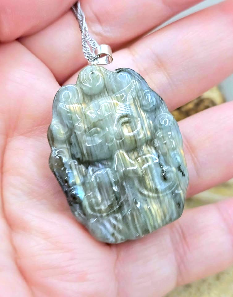 A carved stone pendant with a silver chain, held in a person's hand. The pendant is made of a polished stone with green, gray, and black marbled patterns.