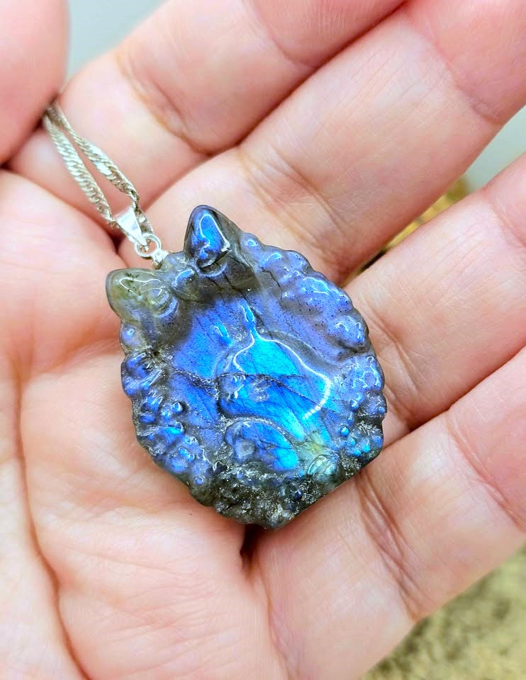 A close-up of a hand holding a rough, irregular-shaped labradorite pendant necklace. The pendant displays vibrant blue and green iridescent flashes with dark gray and black edges, suspended from a silver-toned chain.